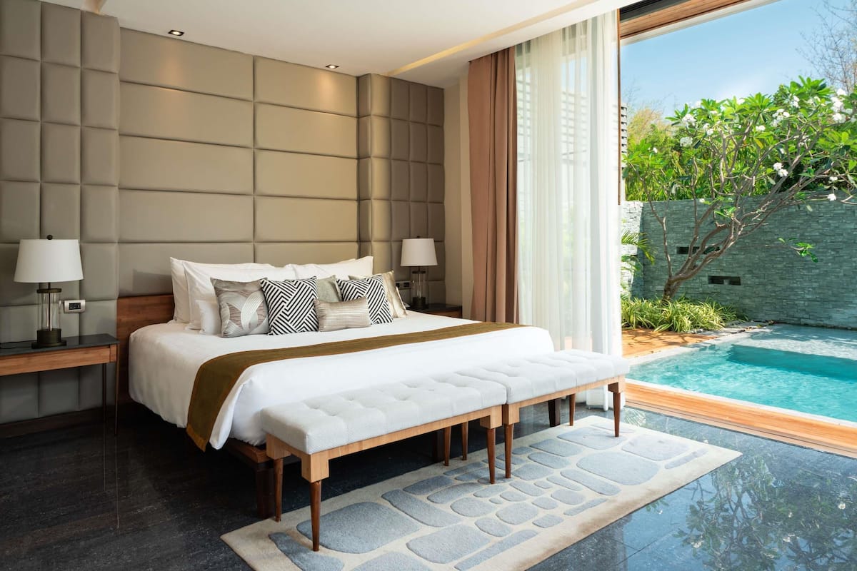 A modern bedroom with a large bed, bedside lamps, a bench, and a view of a swimming pool.