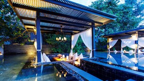 A serene outdoor pool area with a covered seating area, a small waterfall feature, and a lush green backdrop.