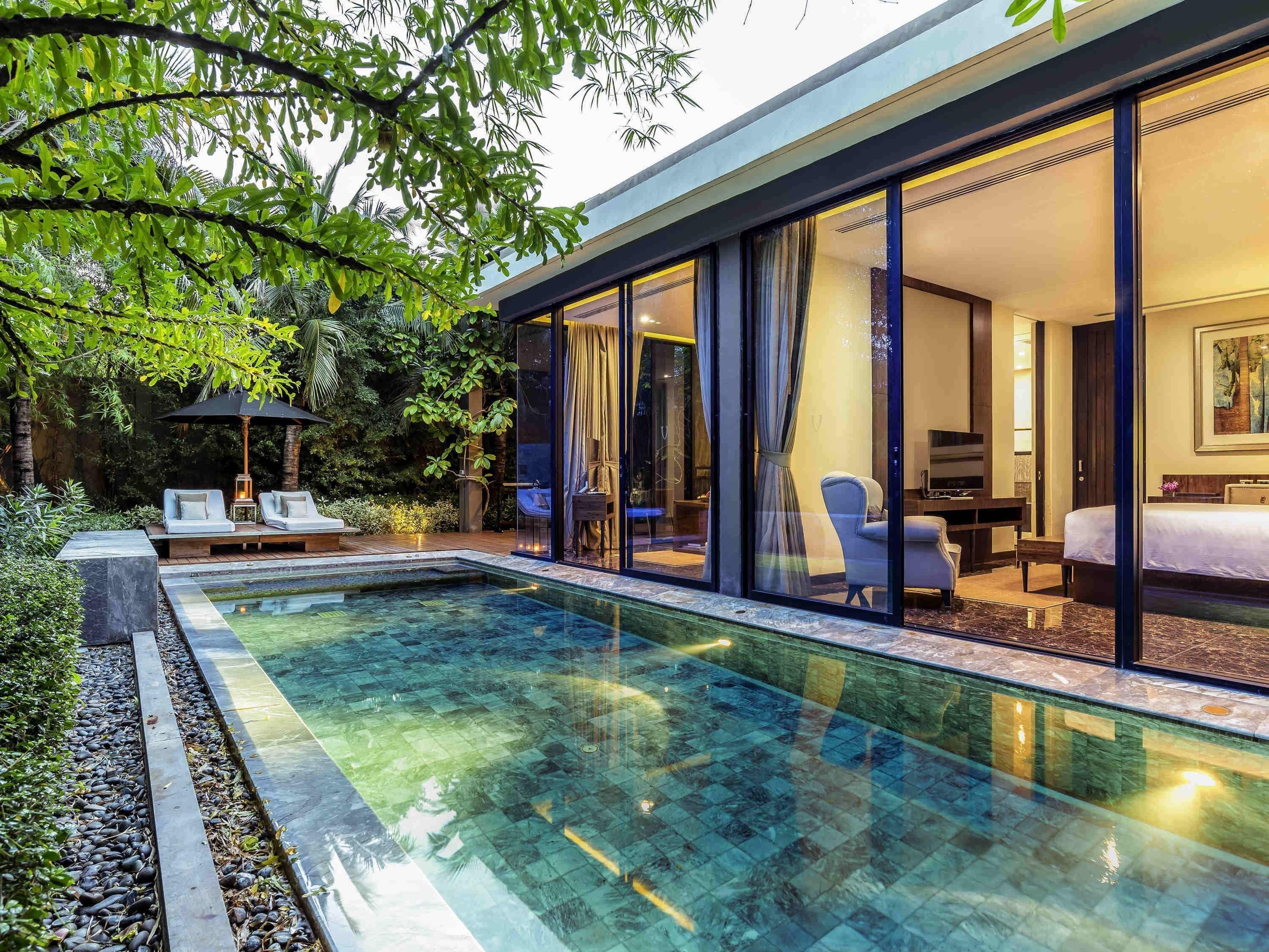 A modern hotel room with a glass wall, a pool, and a seating area with lounge chairs.