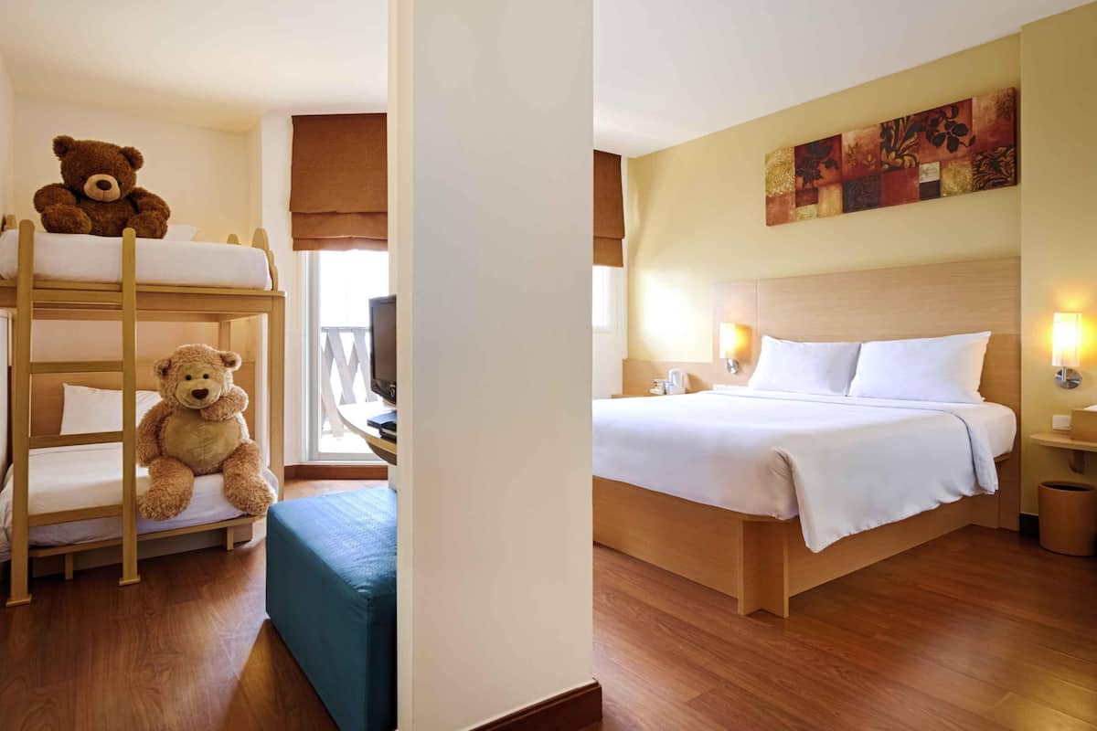 A hotel room with bunk beds, a single bed, a bedside table with a lamp, a painting on the wall, and a window with a view.