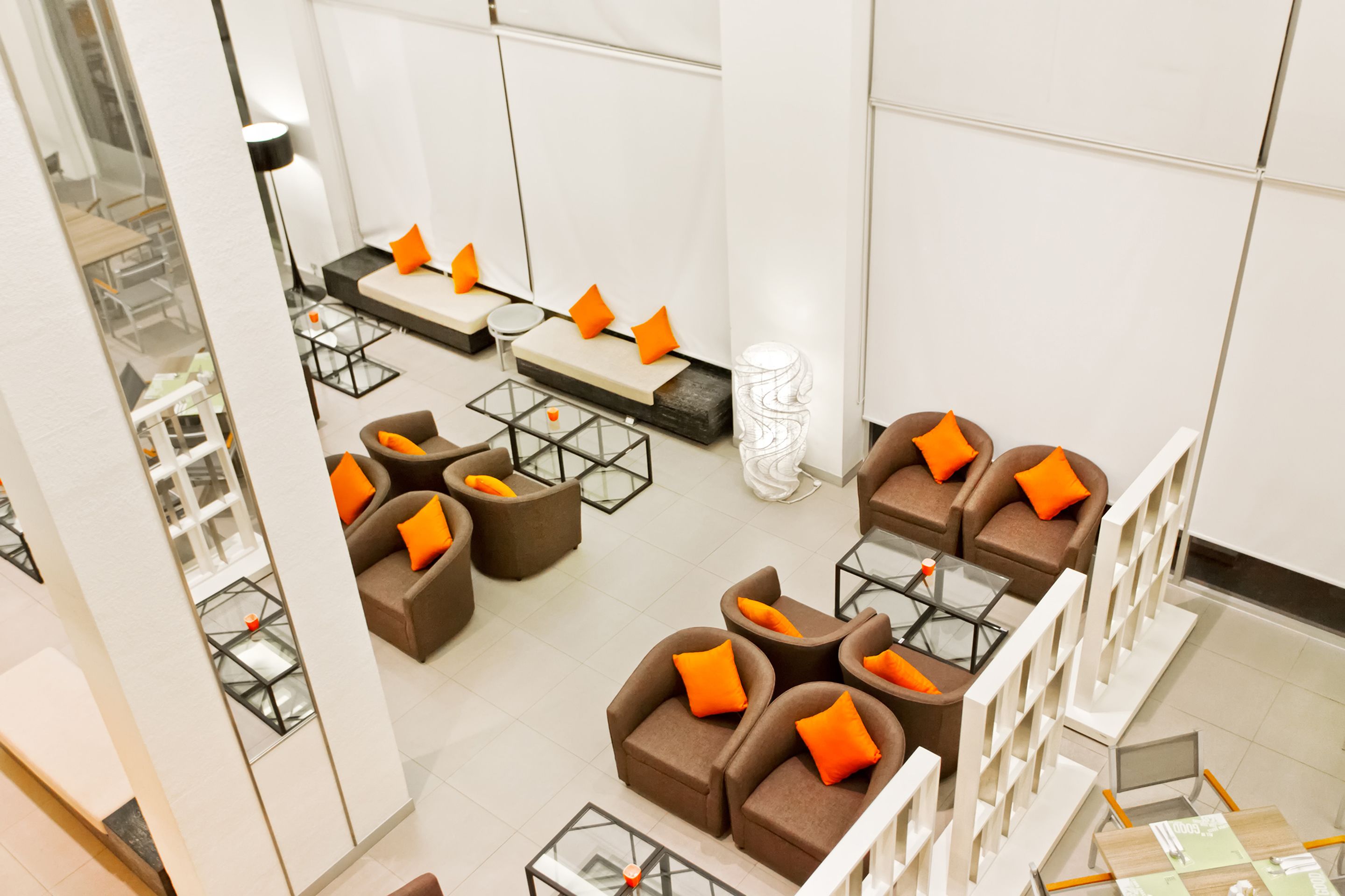 A modern hotel lobby with multiple seating areas, including sofas and armchairs with orange cushions, and a central coffee table.