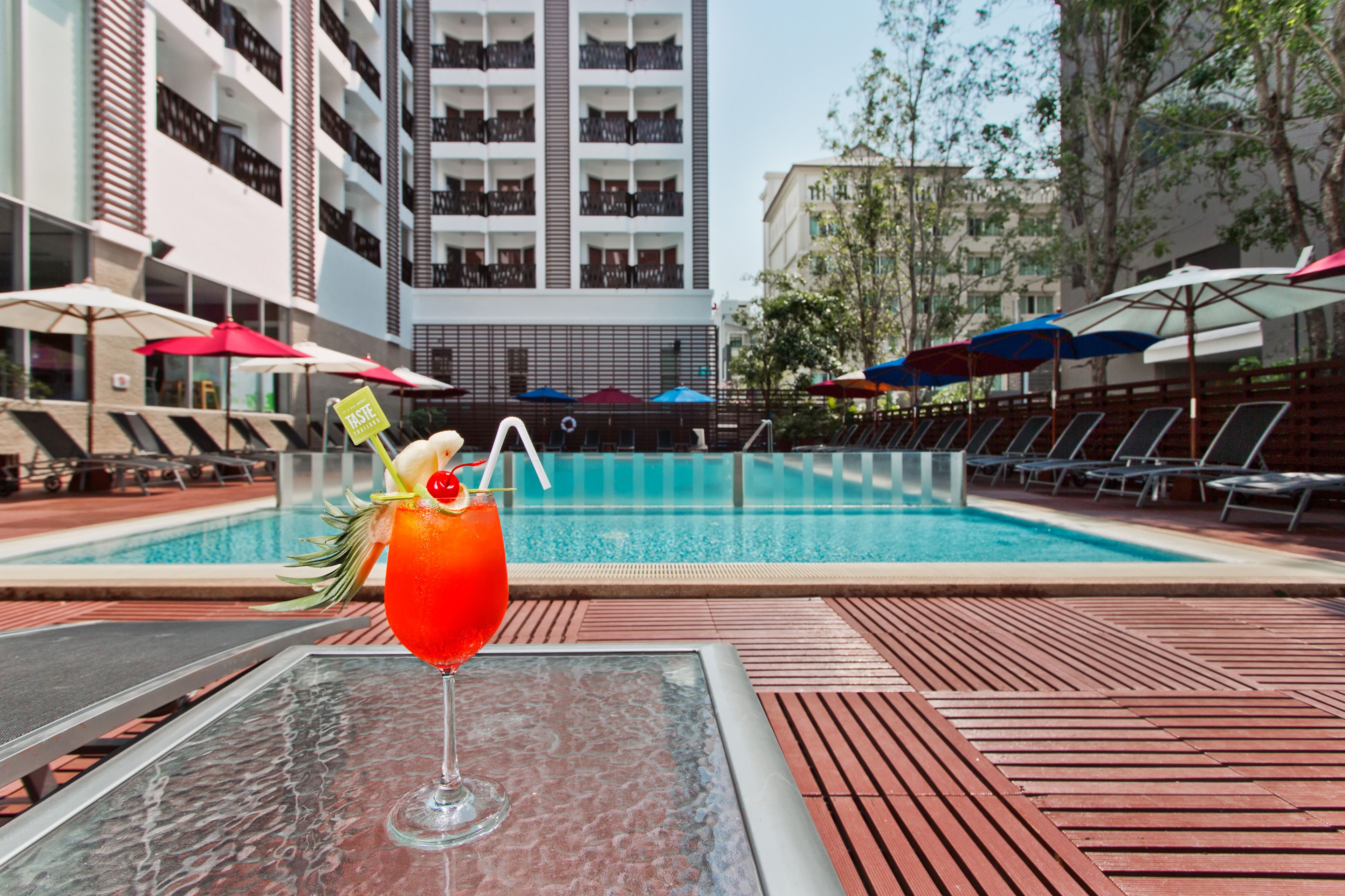A poolside area with a glass table, a drink with a straw and garnish, and lounge chairs with umbrellas.