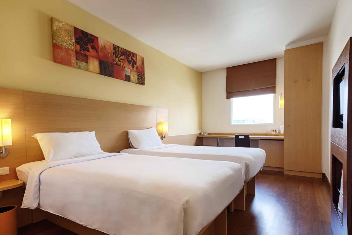 A hotel room with two beds, a wooden headboard, bedside lamps, a window with a view, and a small kitchenette.