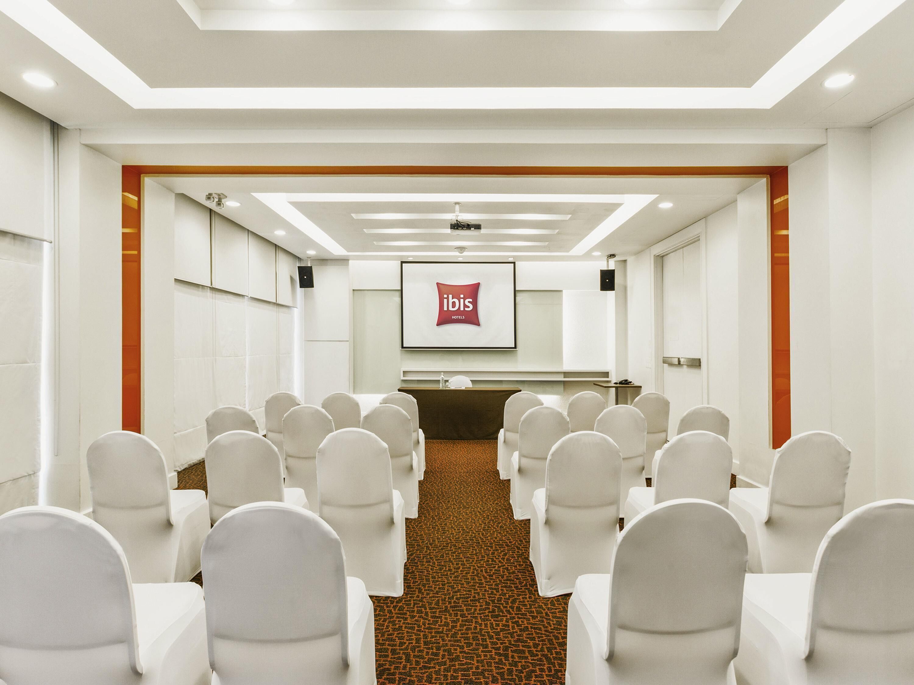 Una sala conferenze con file di sedie bianche, un podio e uno schermo che mostra il logo ibis.