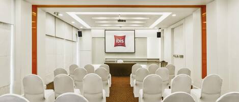 Una sala conferenze con file di sedie bianche, un podio e uno schermo che mostra il logo ibis.