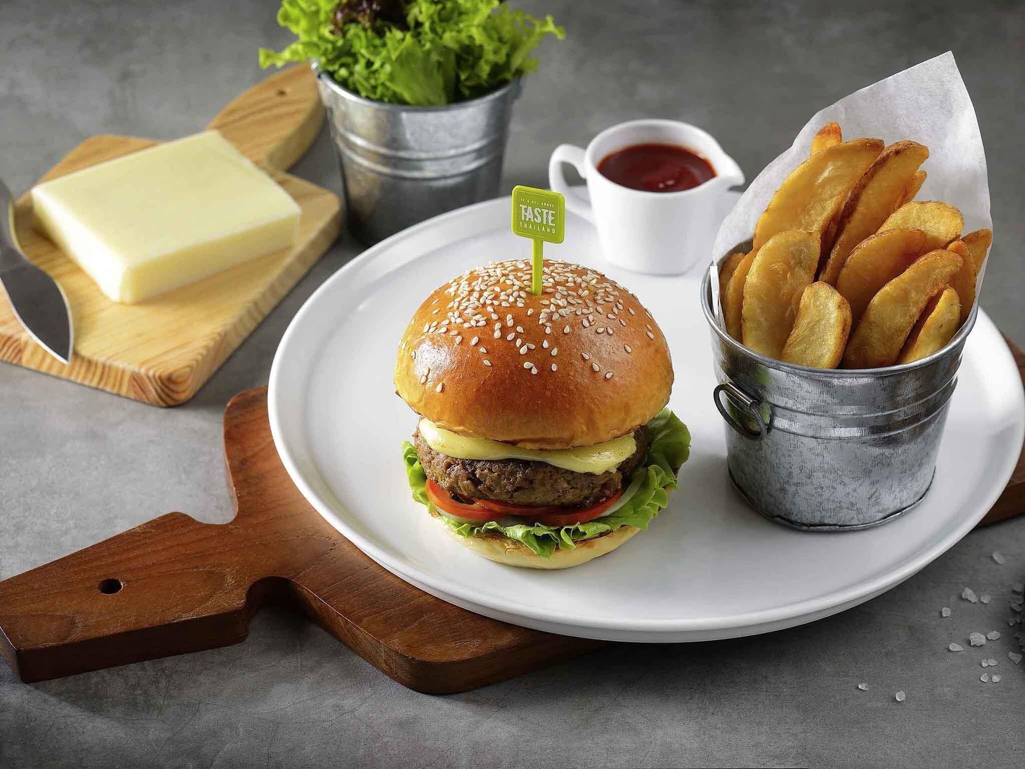 A burger with fries and ketchup on a plate, a knife, a cutting board, and a bucket of fries.