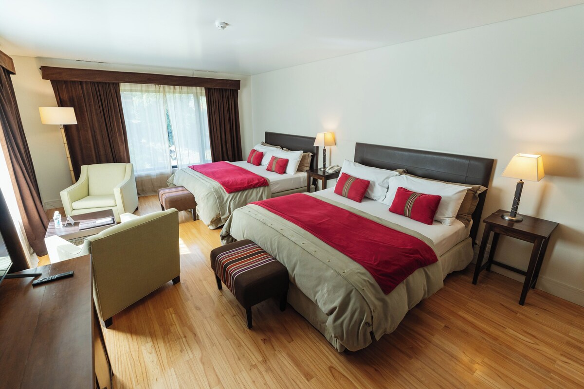 A hotel room with two beds, a chair, a small table, and a window with curtains.