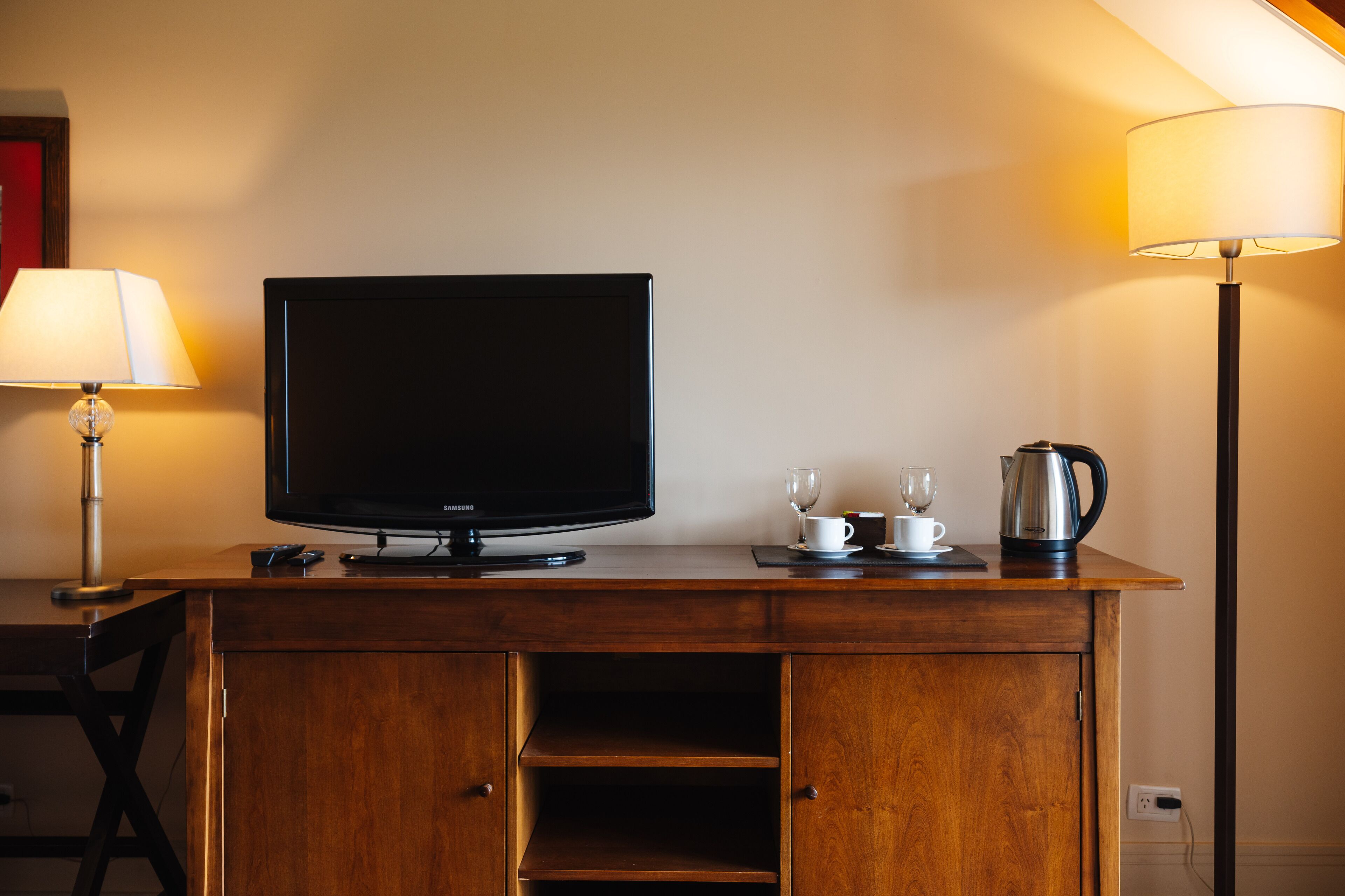 Ein Holztisch mit einem Flachbildfernseher, einer Lampe, einer Tasse, einem Glas und einer Kaffeekanne.