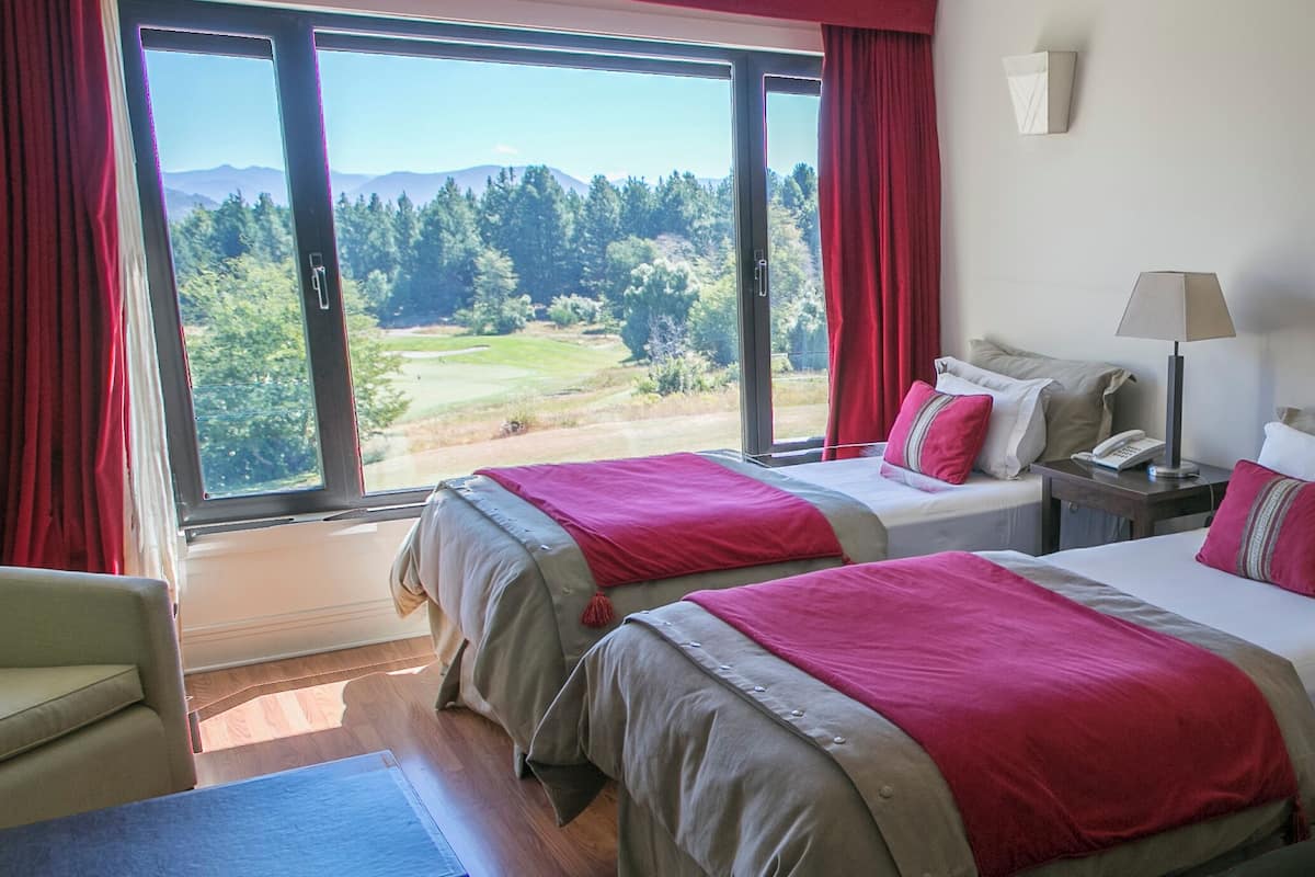 Ein Hotelzimmer mit zwei Betten, einem Sofa, einem Schreibtisch mit Lampe und Blick auf einen Golfplatz durch große Fenster.