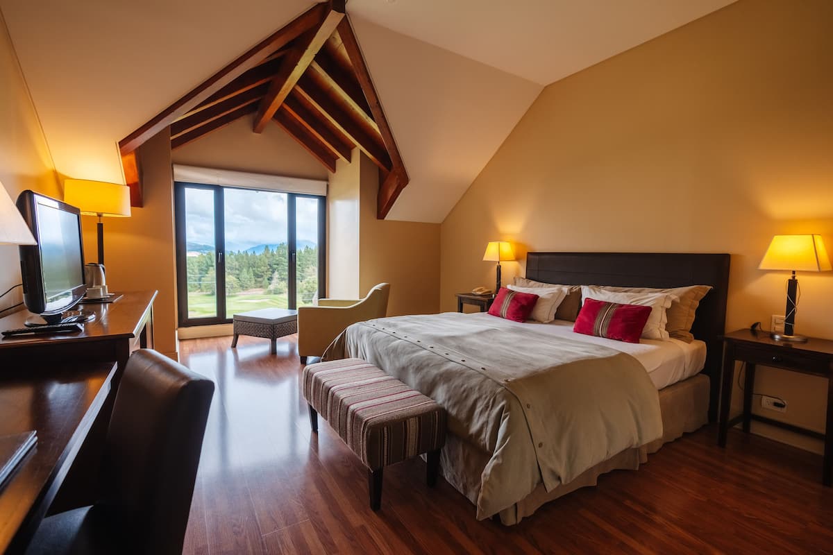 Ein Schlafzimmer mit einem großen Bett, einem Schreibtisch mit Computer, einem Sessel, einer Bank und einem Fenster mit Blick auf Grünfläche.