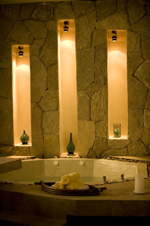 A stone wall with three vertical light fixtures and a bathtub with a towel and a bottle on the counter.