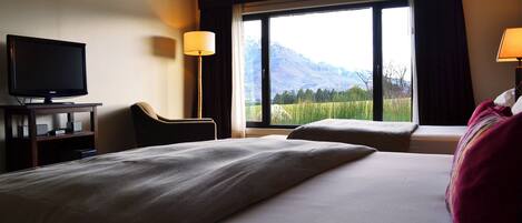A hotel room with a bed, a TV, a chair, and a large window with a mountain view.