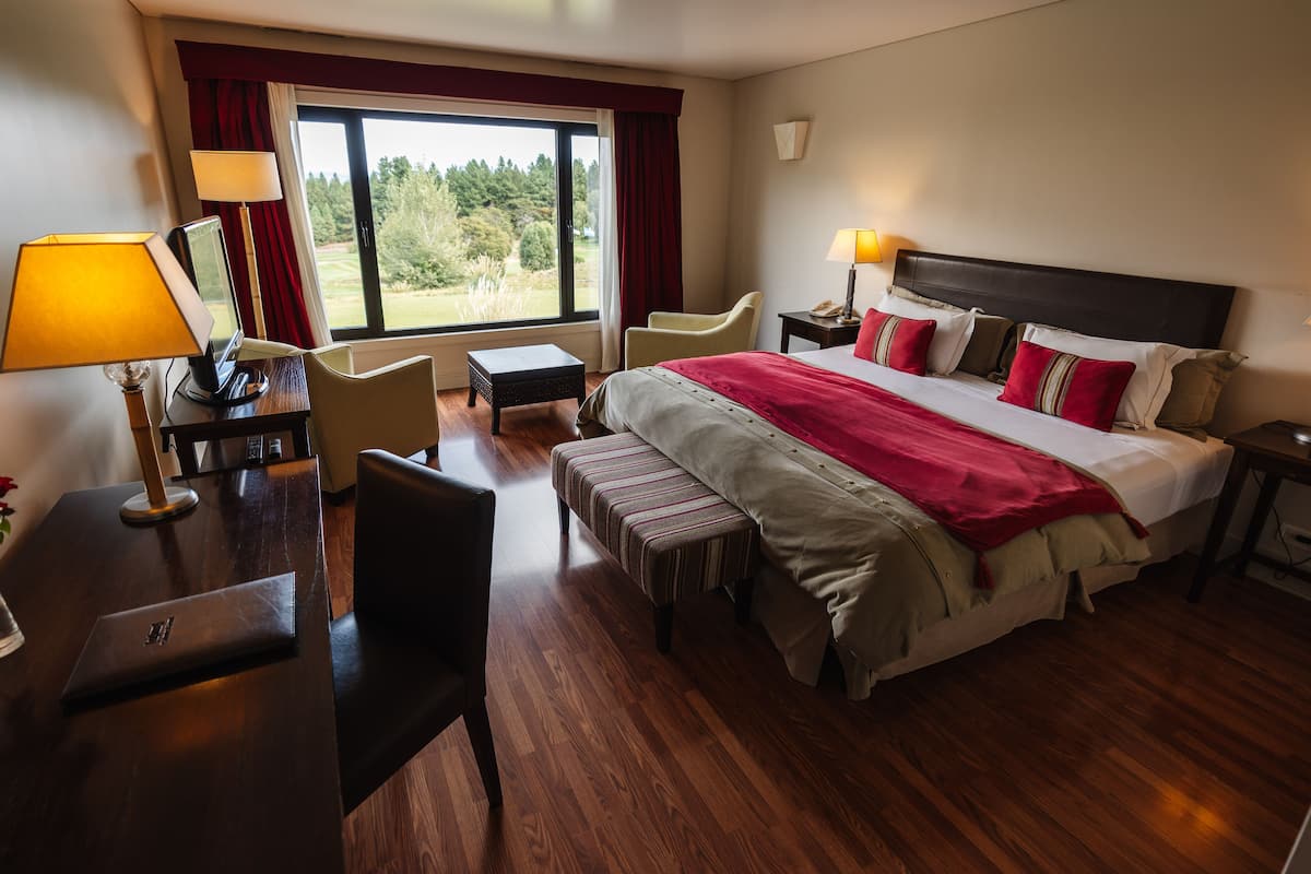 Ein Hotelzimmer mit einem großen Bett, einem Schreibtisch mit Computer, einem Sessel, einem kleinen Tisch und einem Fenster mit Blick auf Grünflächen.