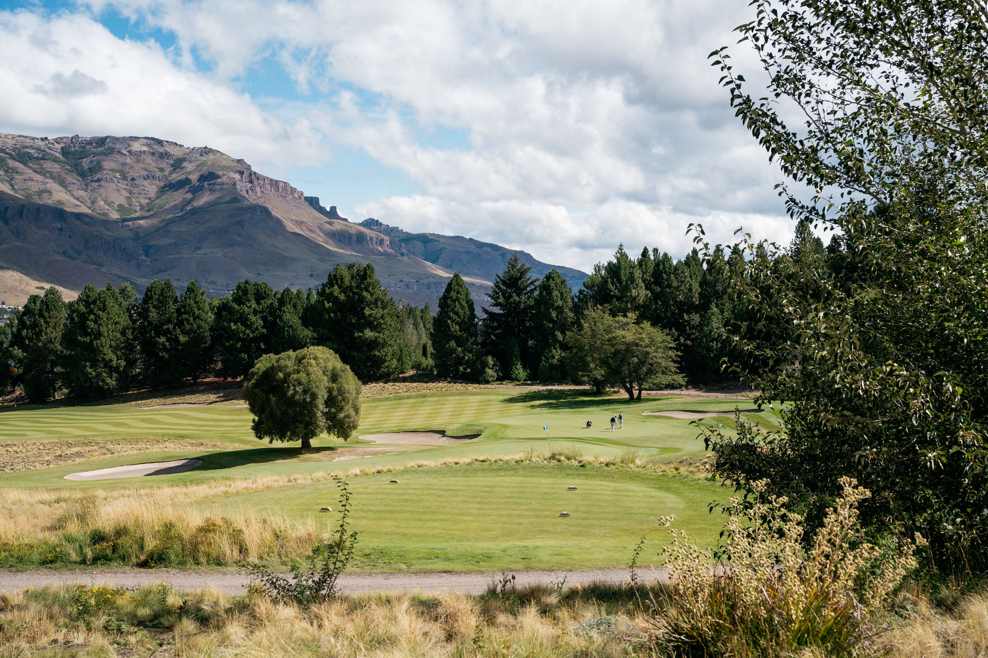 A golf course with a mountainous backdrop, surrounded by dense trees and a clear sky.