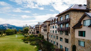 Ein mehrstöckiges Hotel mit Steinvorsprüngen, großen Fenstern und Balkonen, die auf eine gepflegte Rasenfläche und ein kleines Gewässer blicken.
