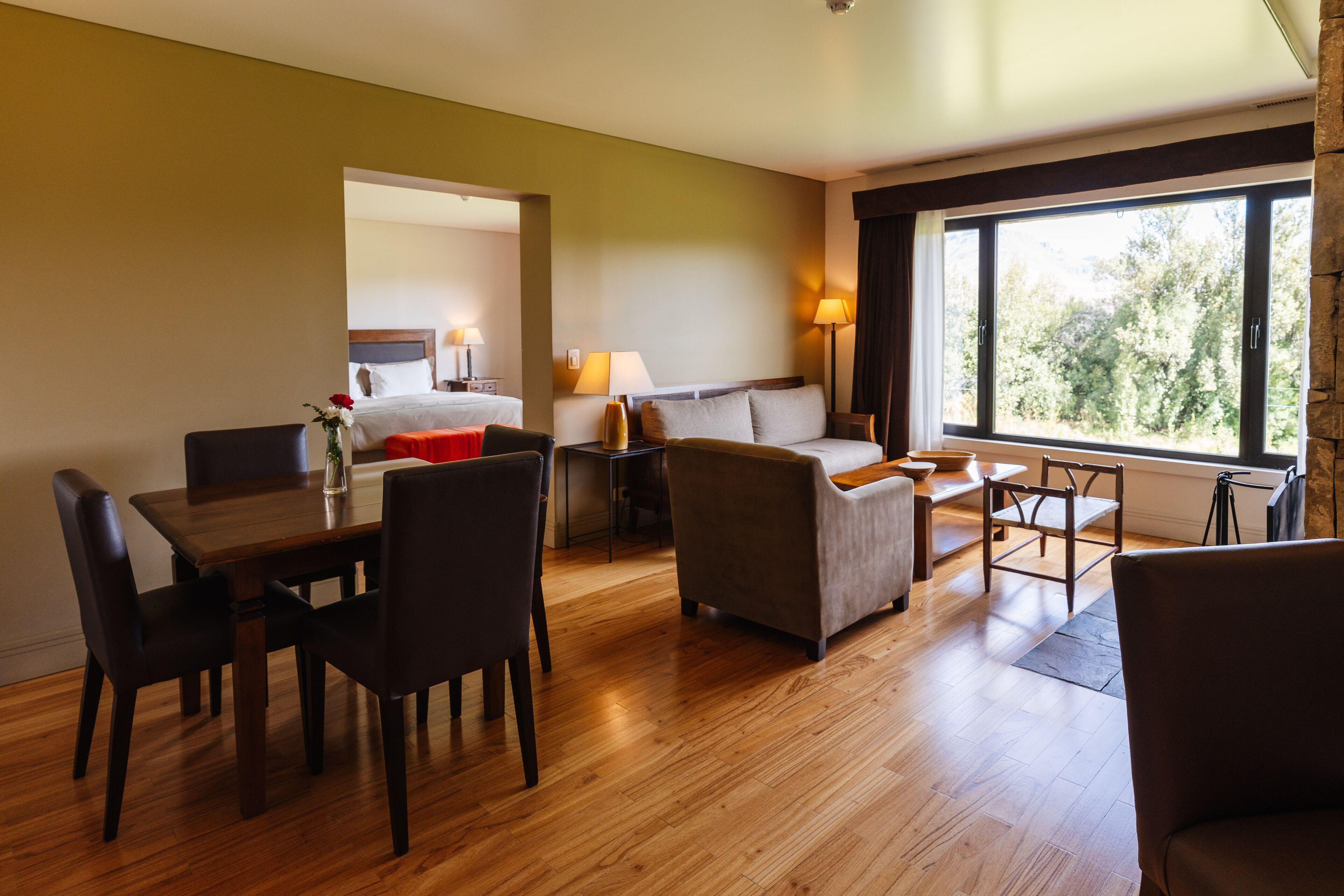 Ein Hotelzimmer mit Holzboden, einem Esstisch mit Stühlen, einer Couch, einem Bett und einem großen Fenster mit Blick auf Bäume.