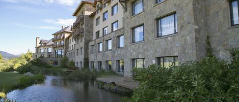 Un hôtel à plusieurs étages, doté d’une façade en pierre et de balcons, avec un étang au premier plan et une chaîne de montagnes en arrière-plan.
