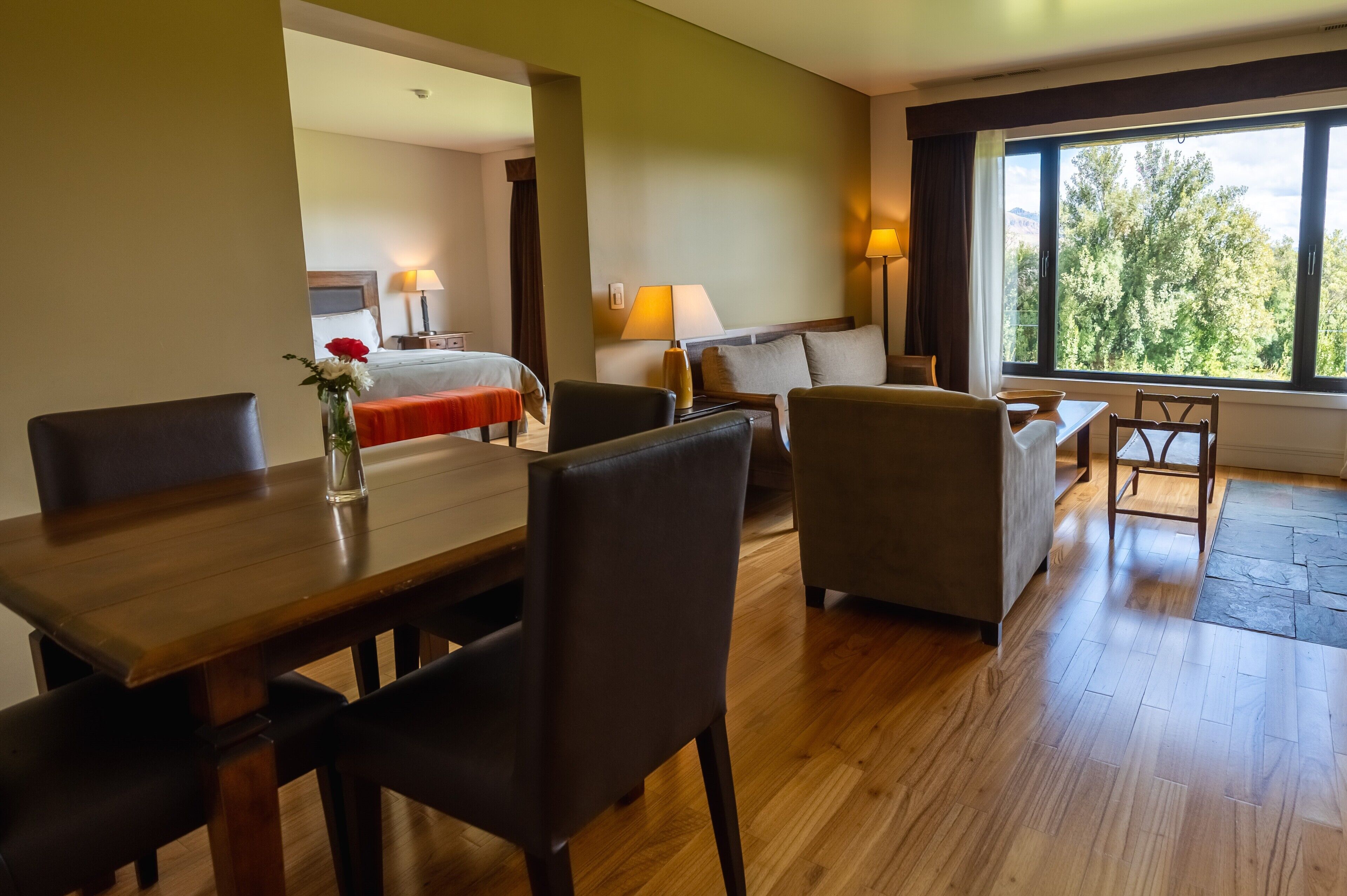 A hotel room with a wooden dining table, two armchairs, a sofa, a bed with a red blanket, and a large window with a view of trees.