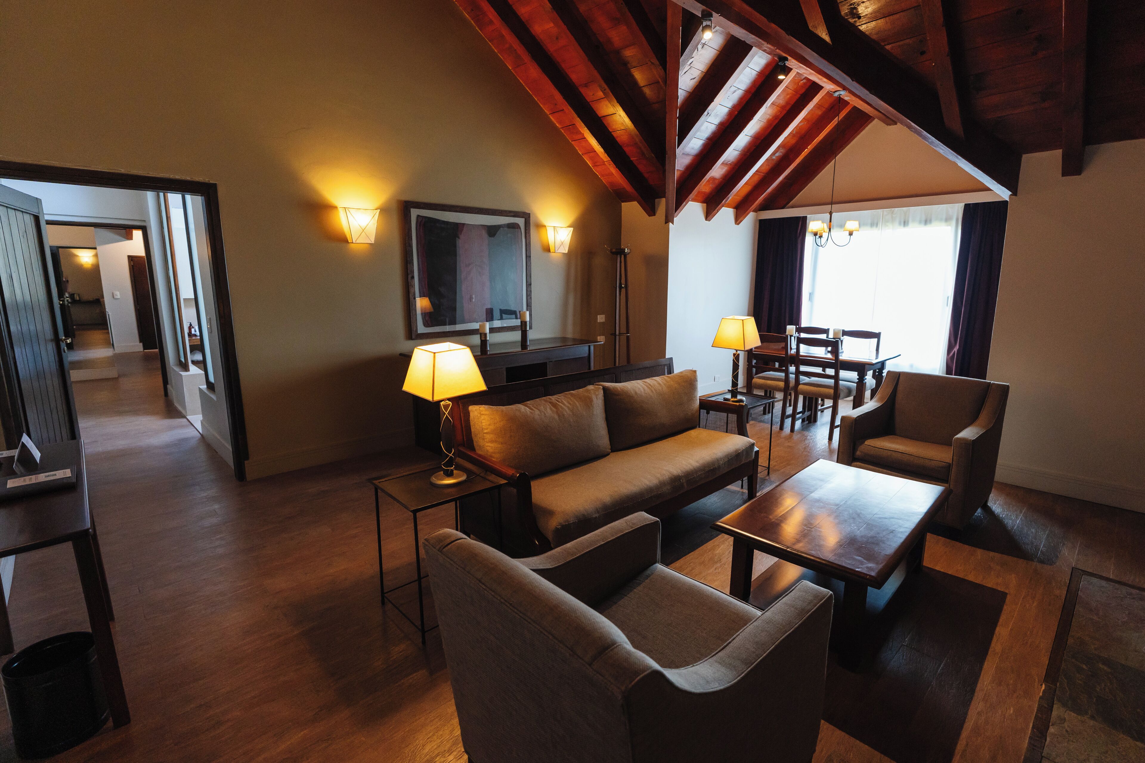 A modern living room with a sofa, armchairs, a coffee table, and a dining table. There are two lamps on side tables, a flat-screen TV, and a large window with curtains.