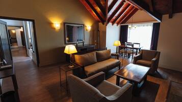 A modern living room with a sofa, armchairs, a coffee table, and a dining table. There are two lamps on side tables, a flat-screen TV, and a large window with curtains.