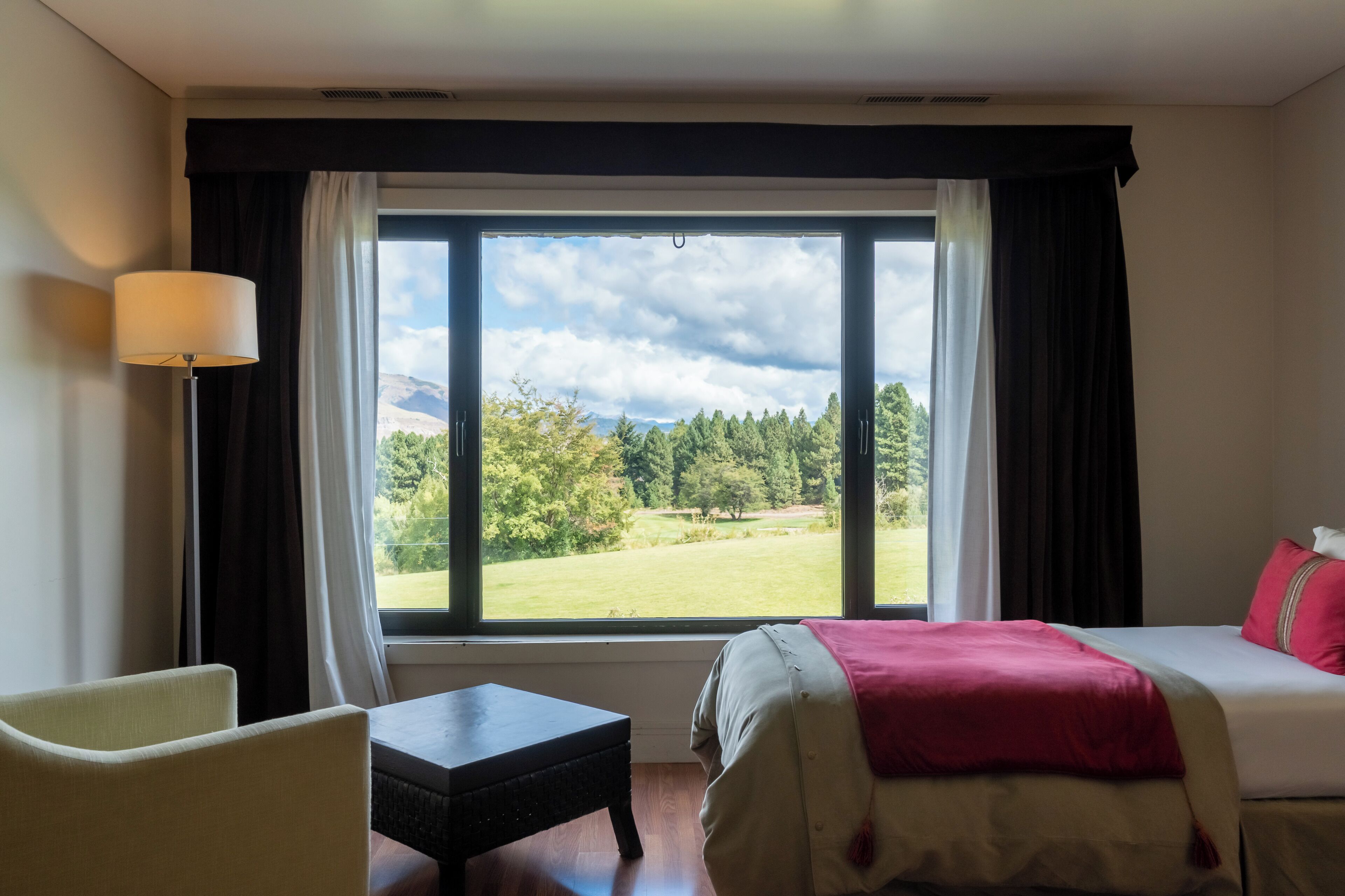 Quarto de hotel com cama, uma cadeira, uma mesinha e uma grande janela com vista para uma paisagem verde e montanhas.