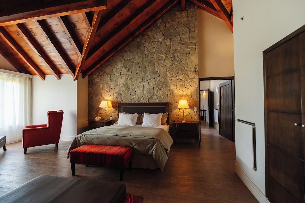 Ein geräumiges Schlafzimmer mit Steinwand, Holzbalken, einem großen Bett, zwei Nachttischen mit Lampen, einem roten Sessel und einer Bank.