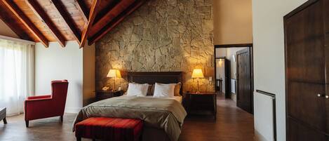 A spacious bedroom with a stone wall, wooden beams, a large bed, two bedside tables with lamps, a red chair, and a bench.