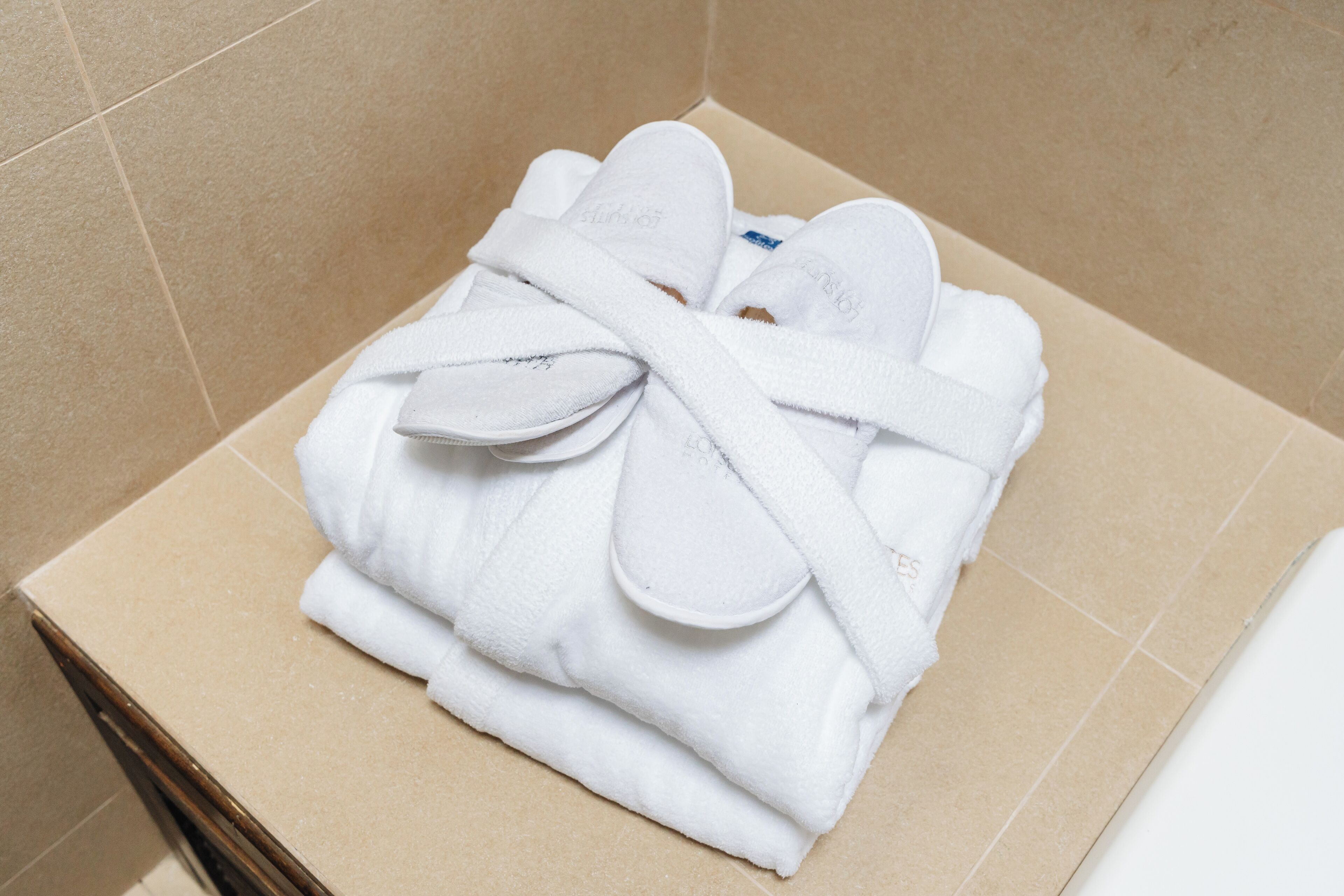Ein ordentlich gefalteter weißer Bademantel und Hausschuhe auf einer braunen Kartonschachtel in einem Badezimmer mit beige gefliesten Wänden.
