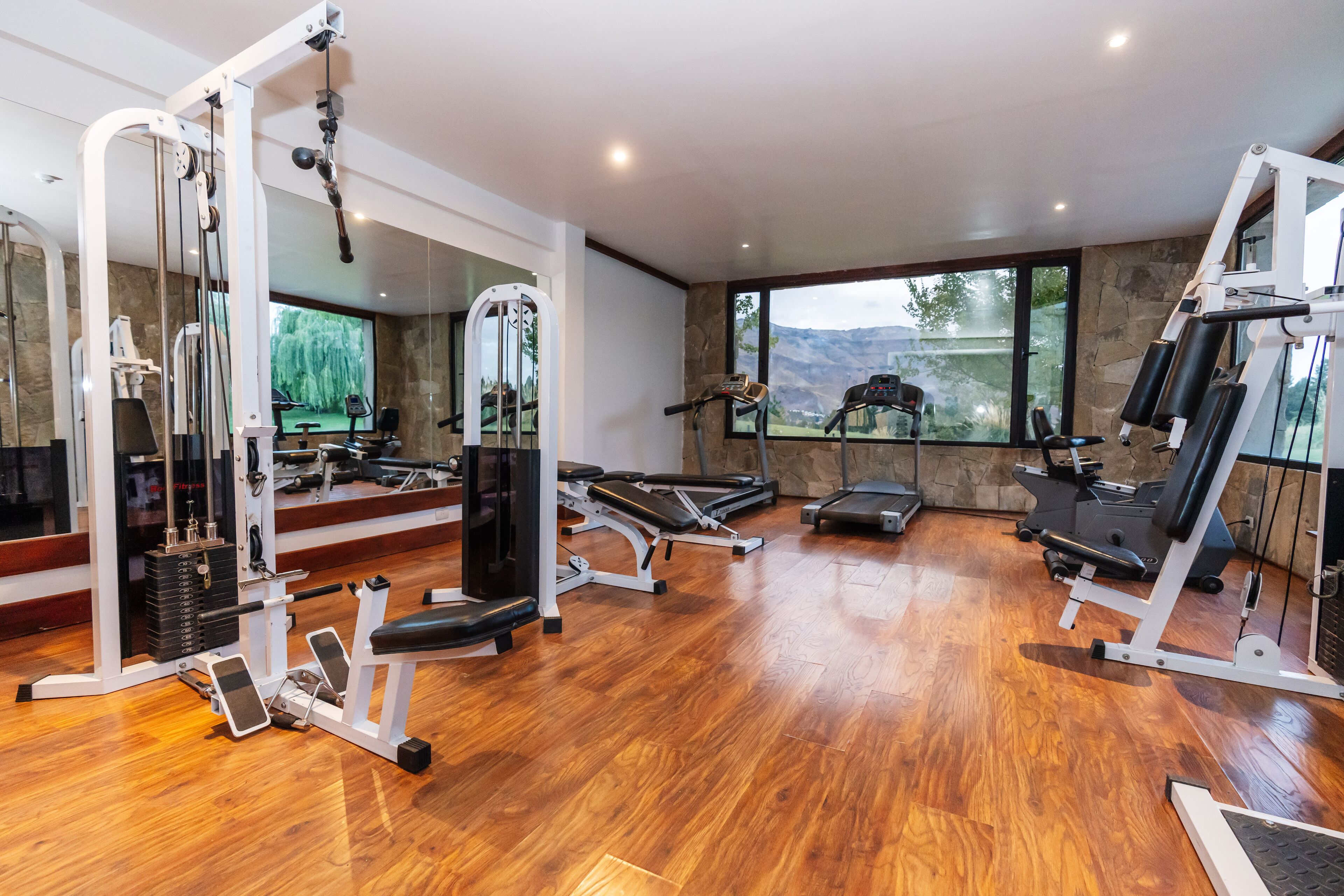 Un gimnasio espacioso con diversos equipos de ejercicio, amplias ventanas que ofrecen vistas al exterior y suelo de madera.