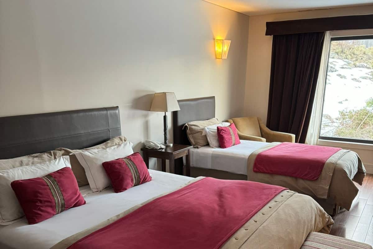 Ein Hotelzimmer mit zwei Betten, jeweils mit Kopfteil, Nachttischen und Lampen. Es gibt ein Fenster mit Vorhängen und Blick ins Freie.
