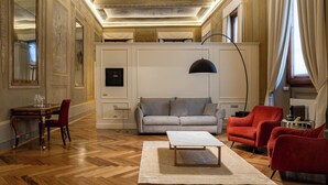 Ein geräumiges Zimmer mit Holzboden, einem grauen Sofa, roten Sessel und einem weißen Couchtisch. Die Decke ist mit aufwendigen Fresken verziert.