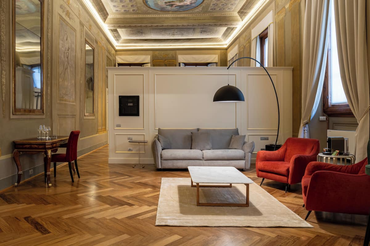 Ruime kamer met een houten parketvloer, een grijze bank, rode fauteuils, een witte salontafel en een plafond met ingewikkelde fresco's.