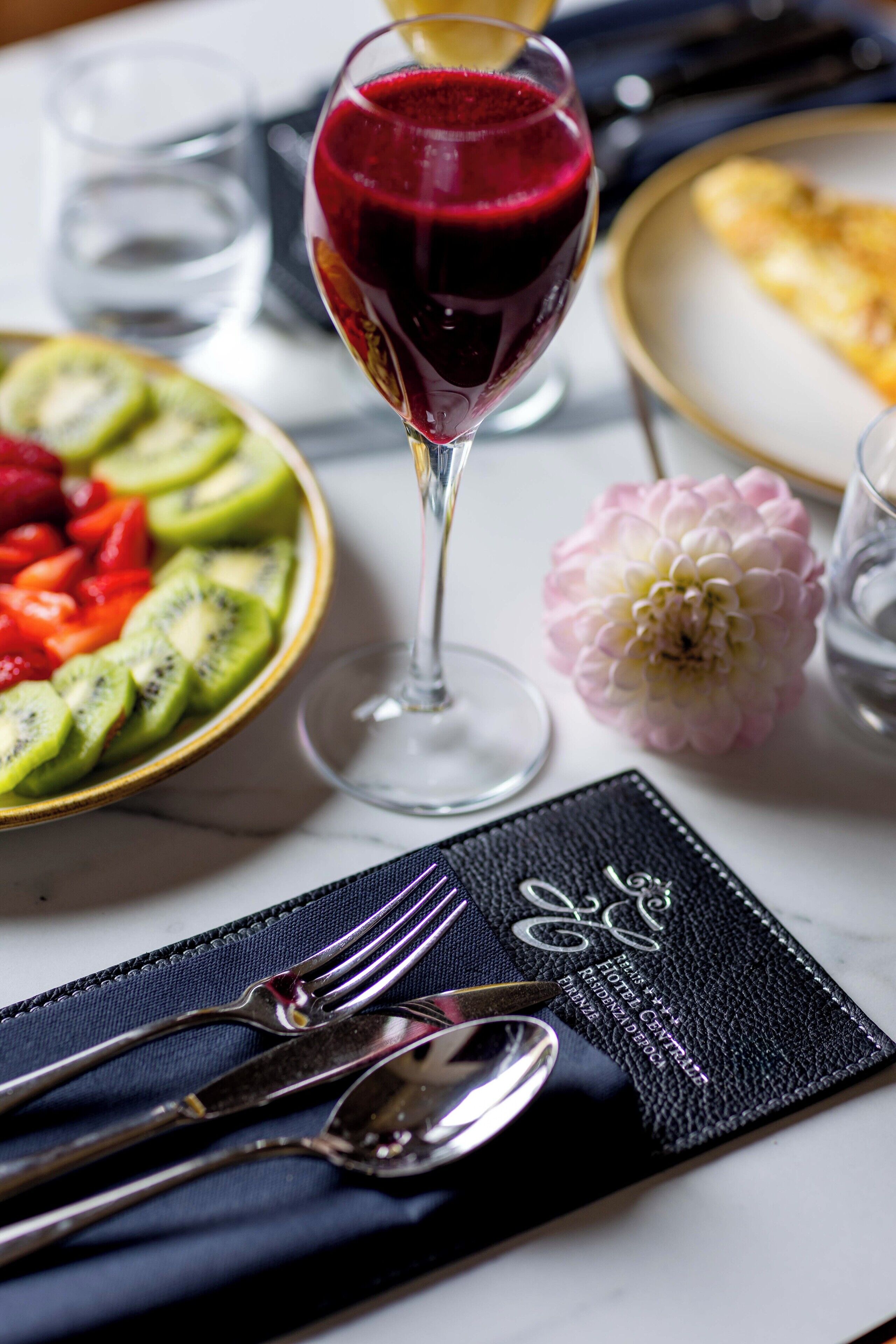 Una mesa de comedor con un vaso de jugo de frutos rojos, un bol de fruta cortada y un plato de comida.