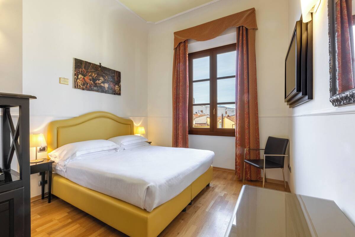 Een hotelkamer met een bed, een bureau met lamp, een stoel, een raam met gordijnen en een schilderij aan de muur.