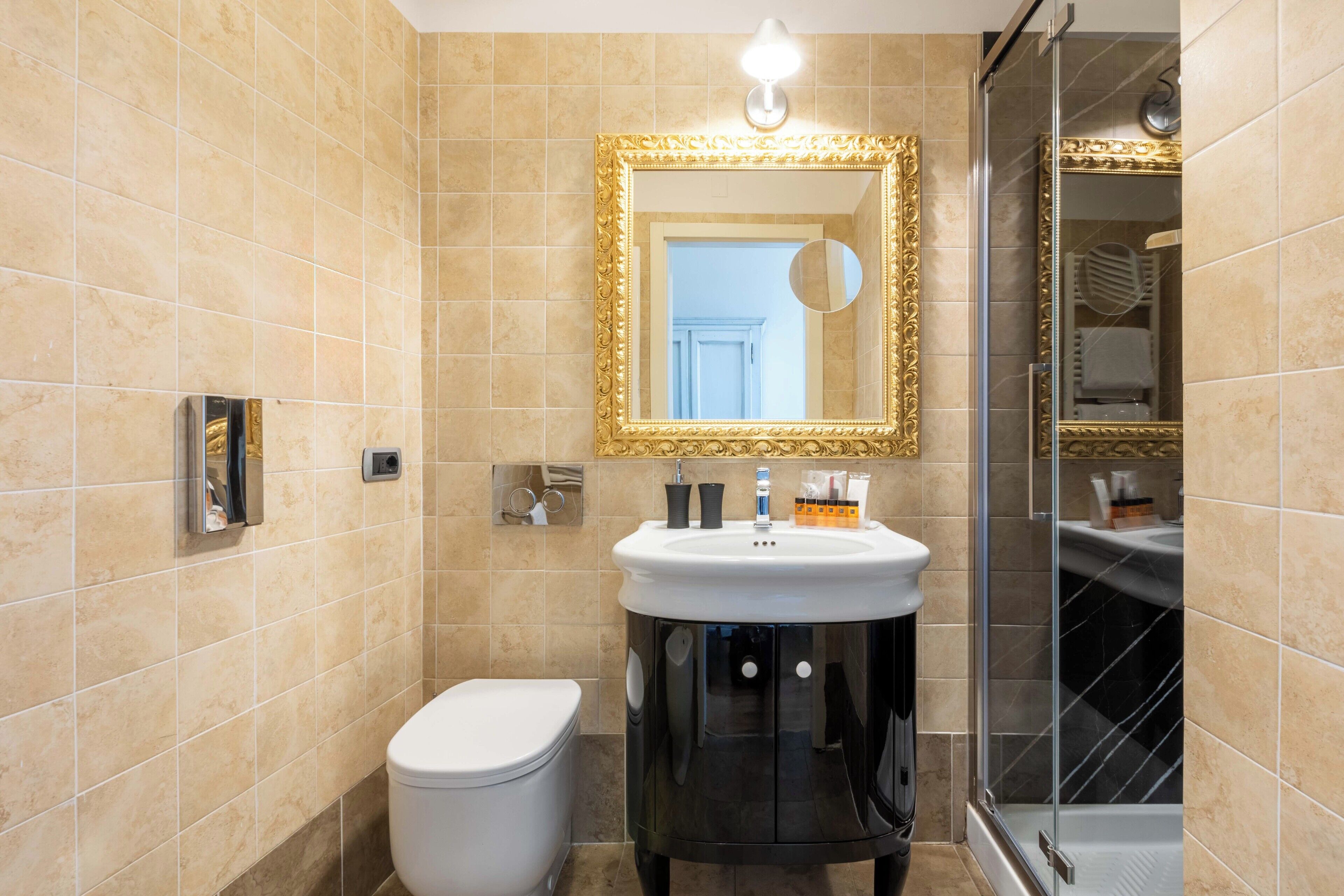 A bathroom with beige tiles, a white toilet, a black vanity with a gold-framed mirror, and a glass shower enclosure.