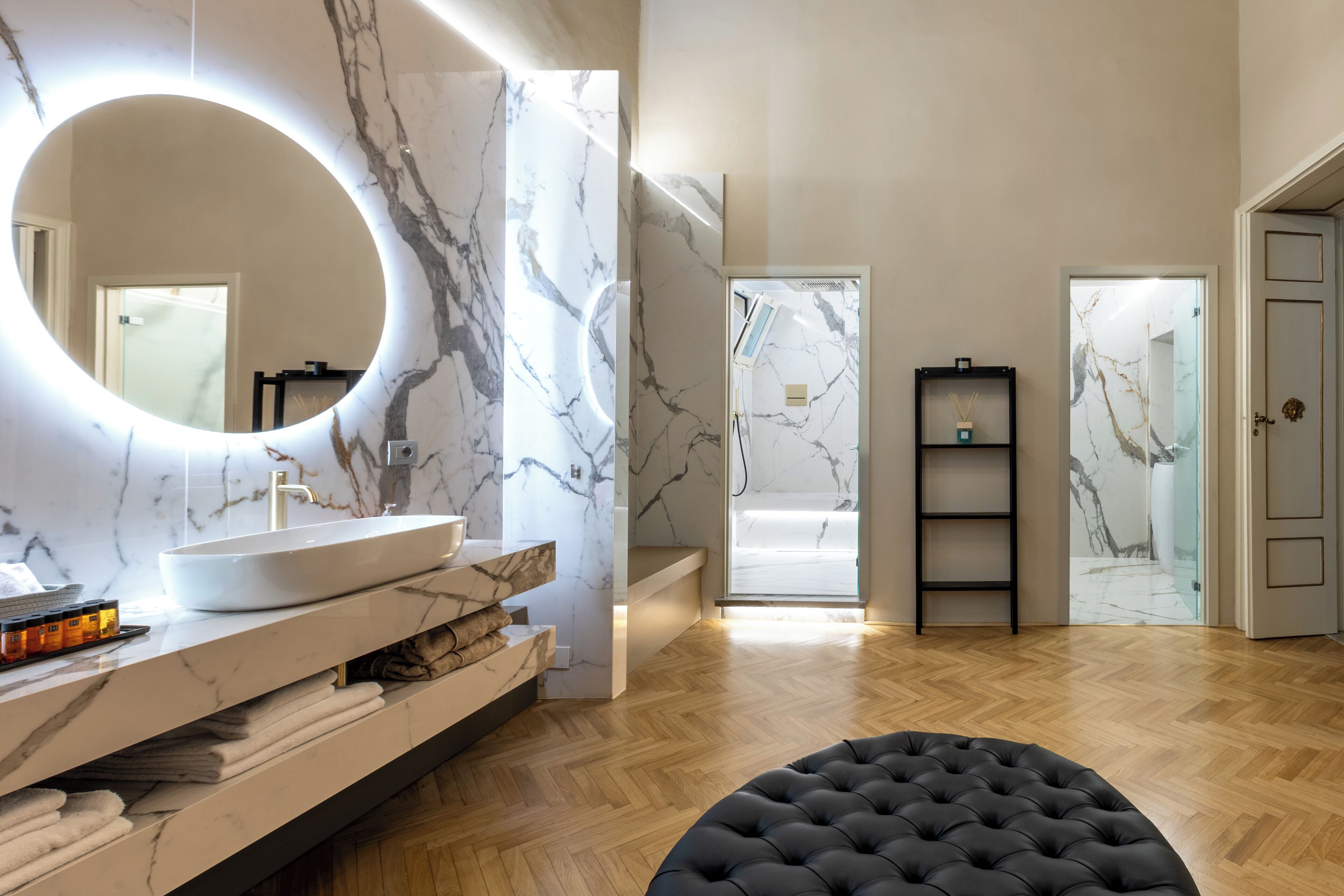 A modern bathroom with a large oval mirror, marble walls, a white sink, and a black ottoman.