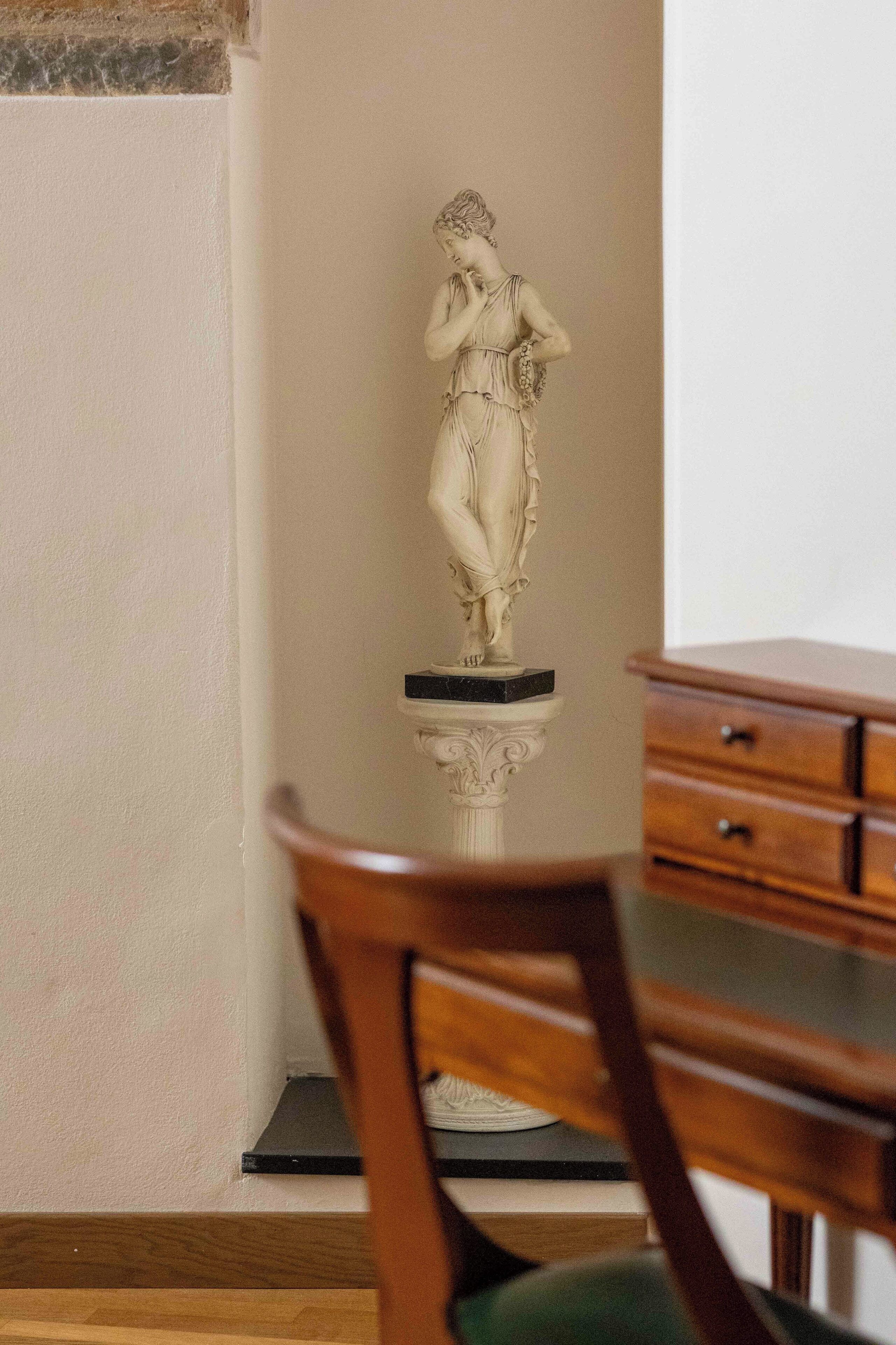 A statue on a pedestal in a room with a wooden chair and a wooden cabinet.