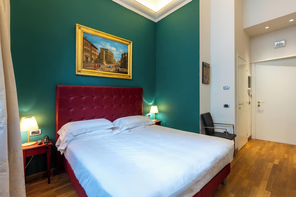 Een hotelkamer met een rood hoofdeinde, een bed met witte lakens, een schilderij aan de muur en een bureau met een lamp.