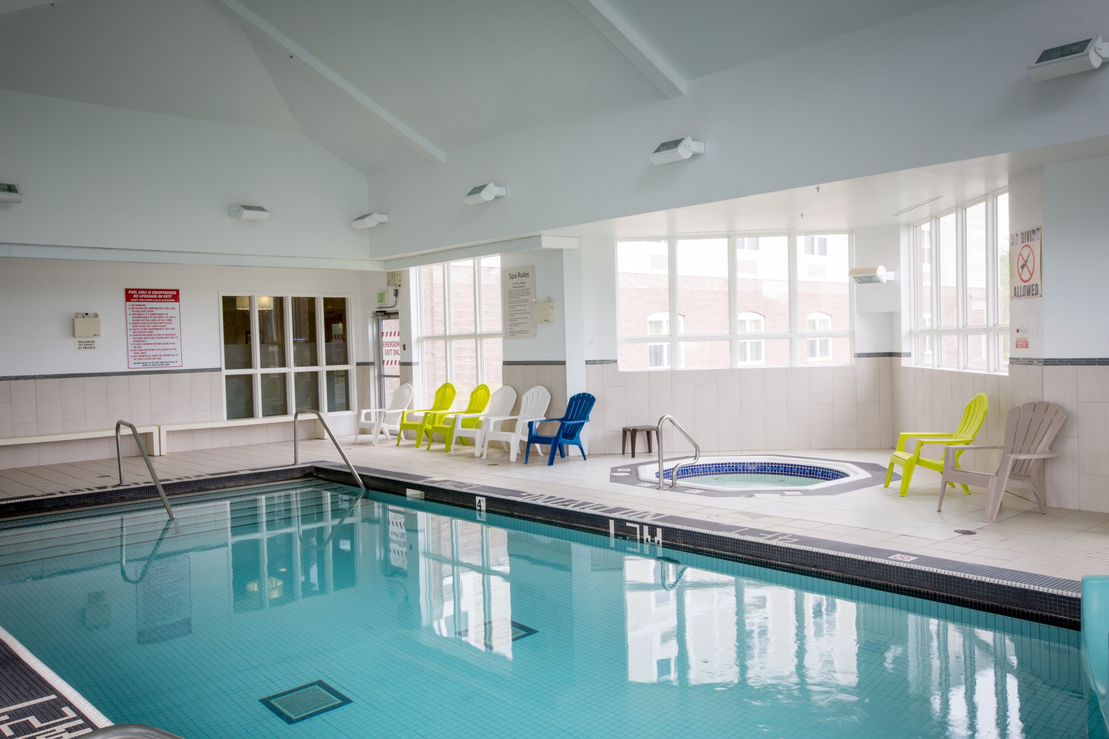 An indoor swimming pool with clear water, surrounded by white walls and windows. There are lounge chairs and a hot tub area.