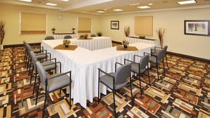 Un salón de conferencias con una gran mesa rectangular rodeada de sillas, una alfombra estampada y cuadros en las paredes.