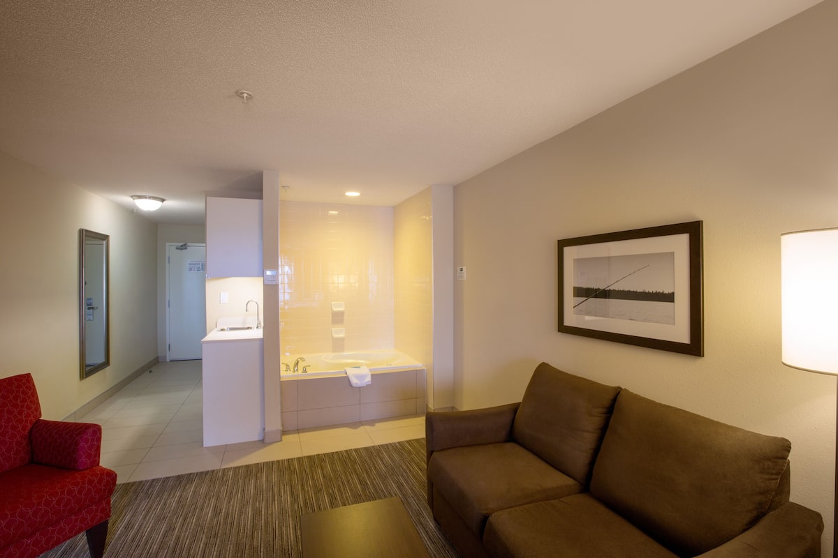 A hotel room with a brown sofa, a red armchair, a small table, a framed picture on the wall, and a bathroom with a sink and bathtub.