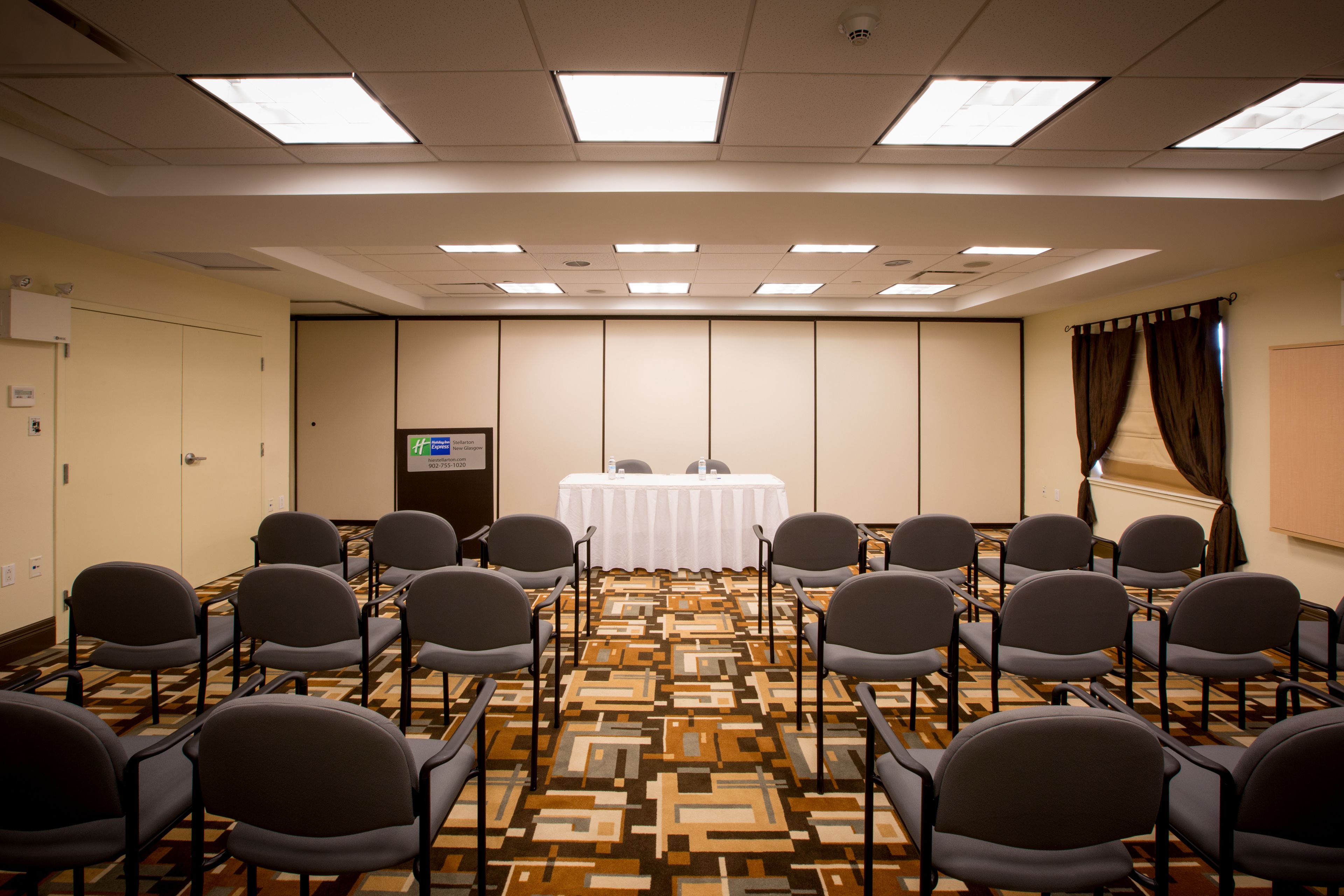Un salón de conferencias con alfombra estampada, filas de sillas, una mesa blanca con pantalla de proyector y una puerta a la izquierda.