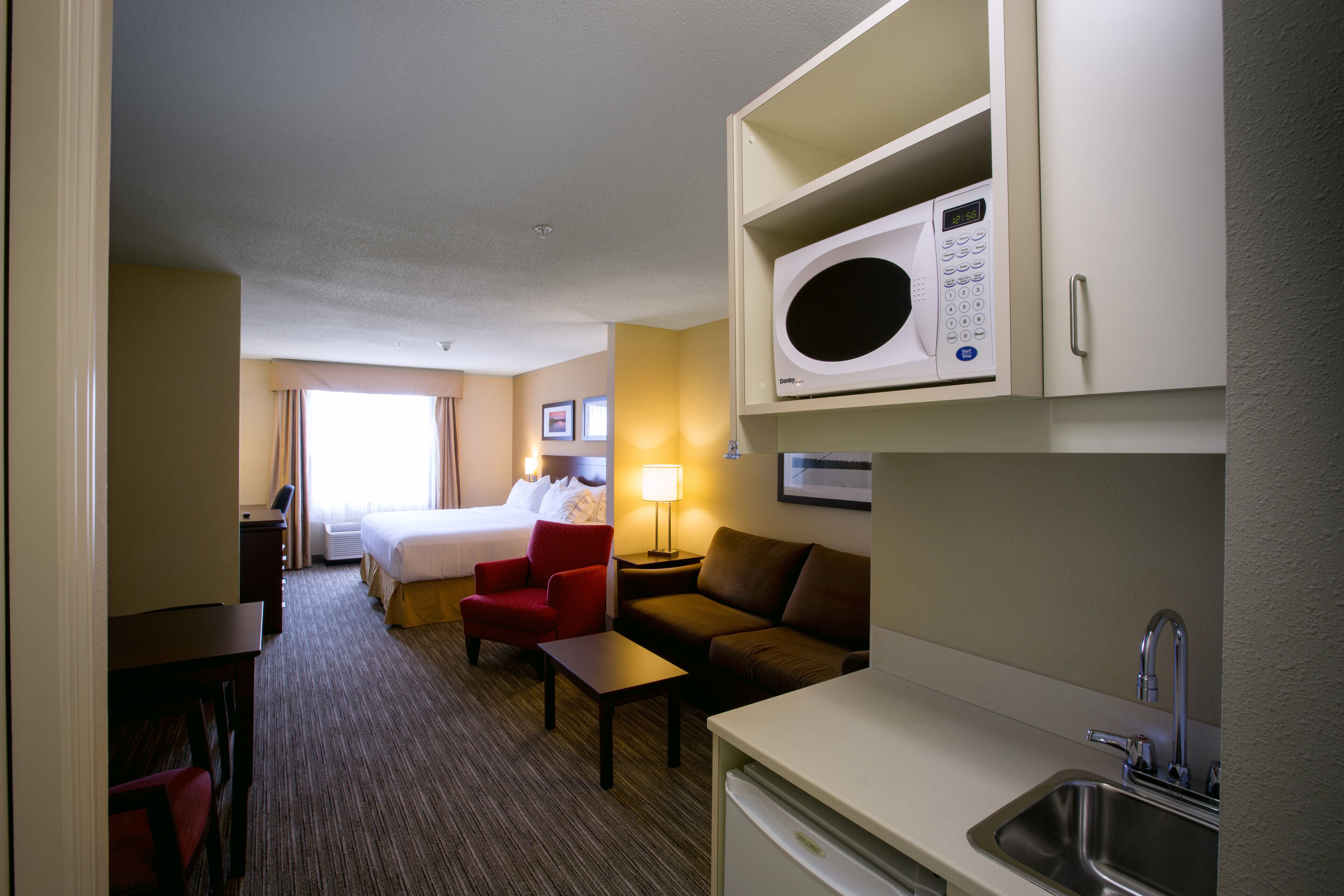 Une chambre d’hôtel avec un lit, un fauteuil rouge, un canapé marron, une petite table, une lampe, un four à micro-ondes et un lavabo.