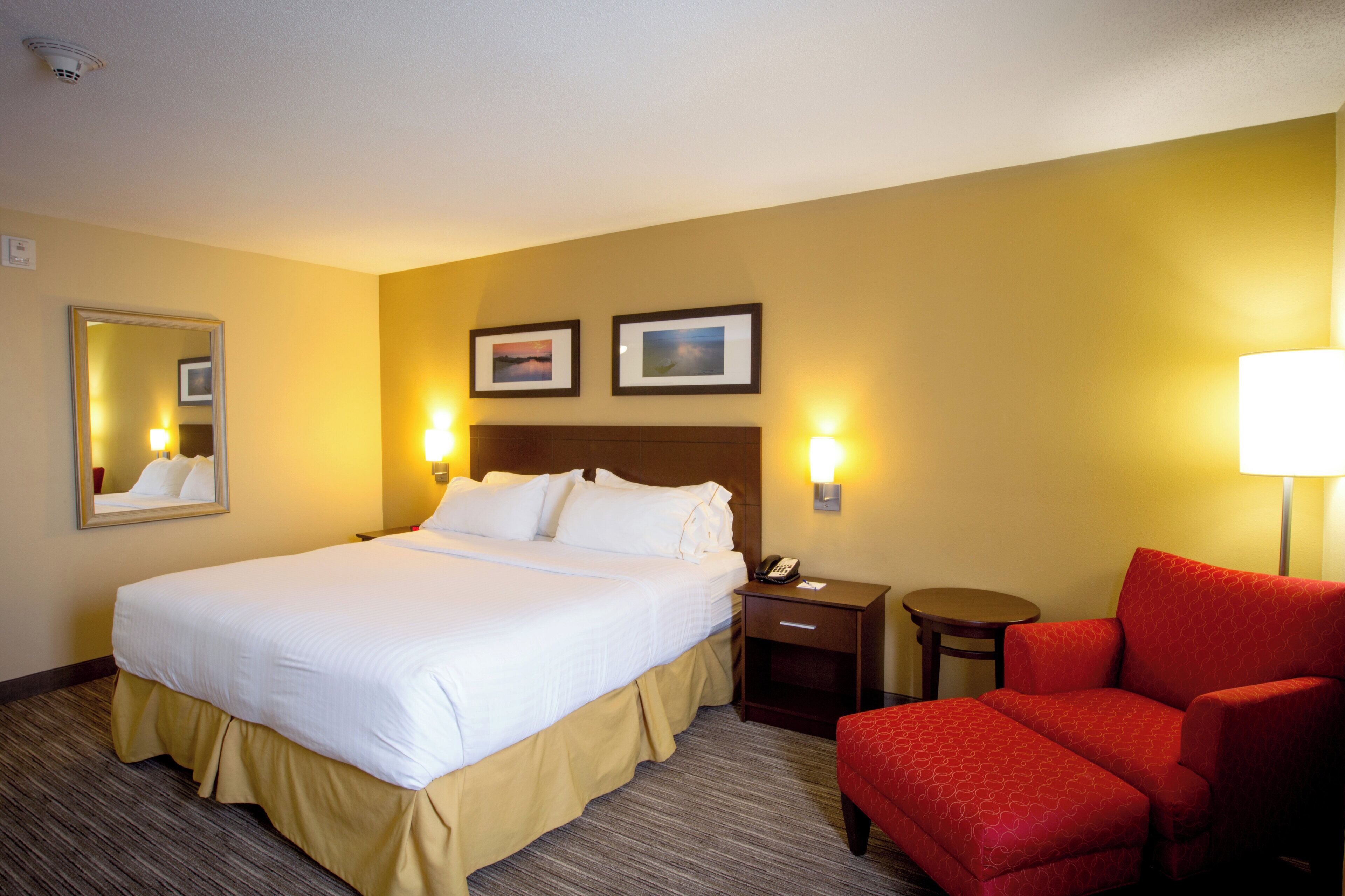 A hotel room with a large bed, a bedside table with a lamp, a chair, and two framed pictures on the wall.