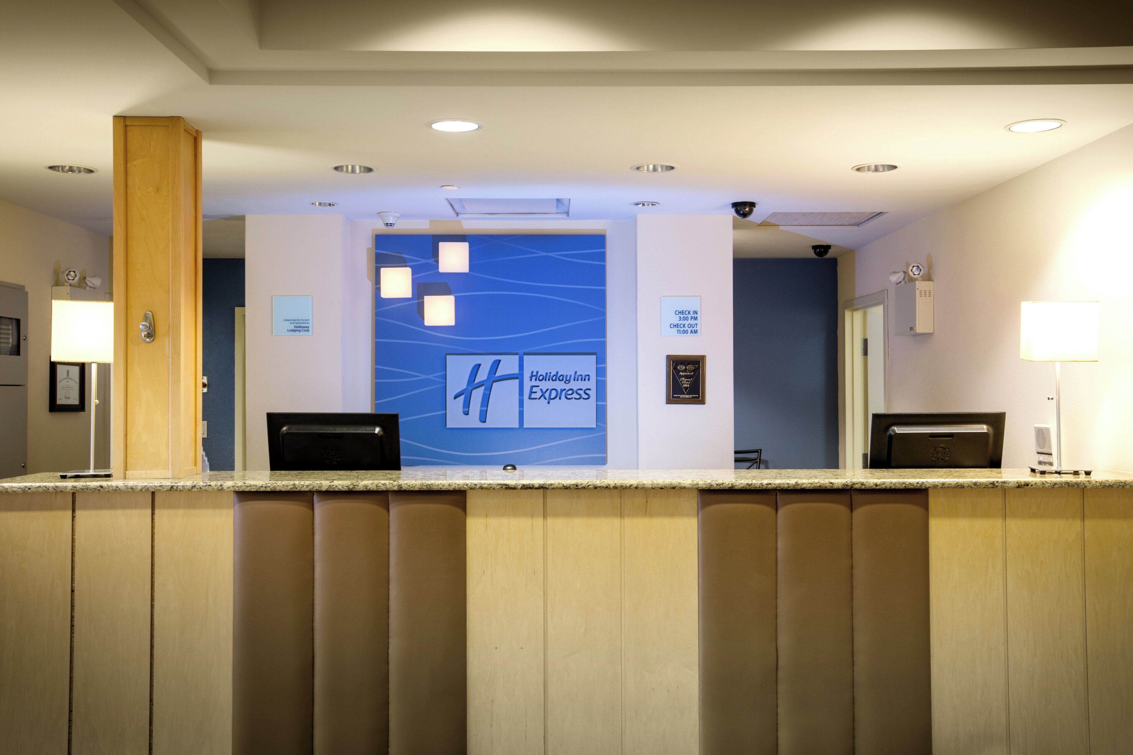 A hotel reception desk with a blue wall featuring the Holiday Inn Express logo. There are two receptionists' stations, a security camera, and a sign indicating check-in and check-out times.