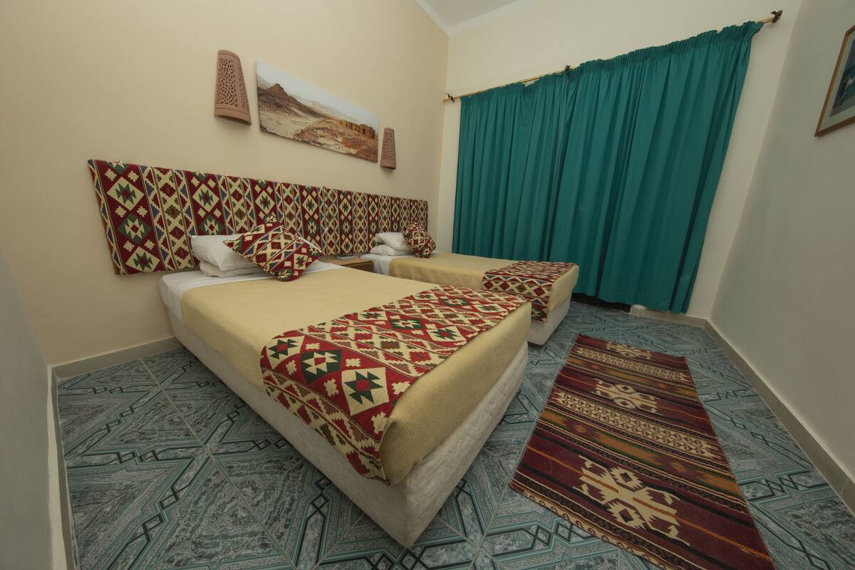 Une chambre d’hôtel avec deux lits, une tête de lit à motifs, une grande fenêtre avec des rideaux de couleur turquoise et un tapis décoratif.