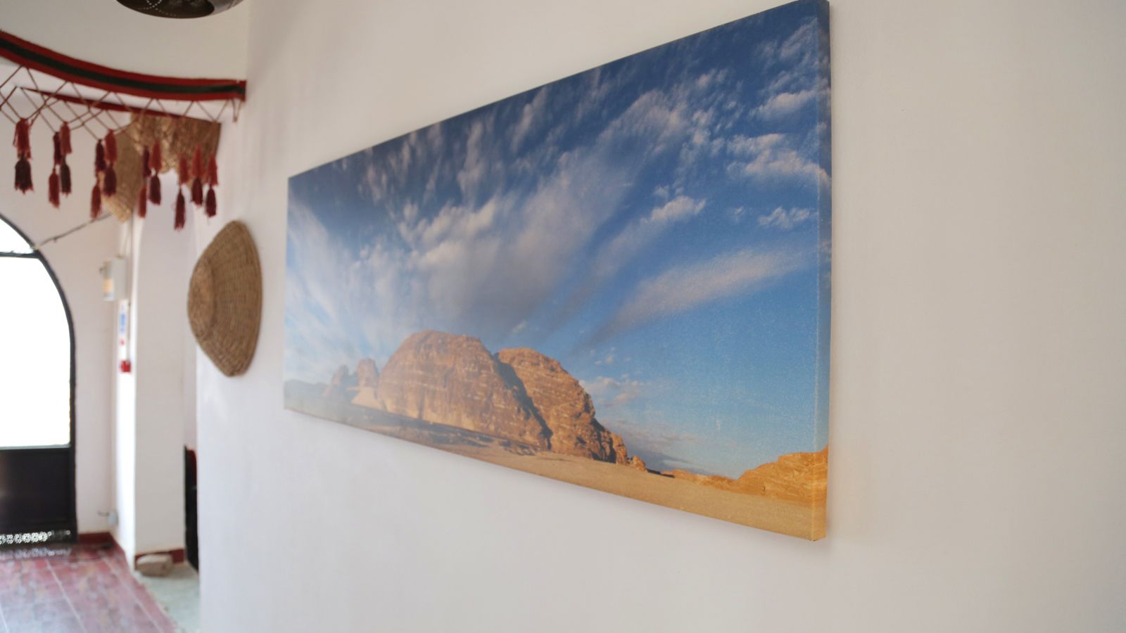 Una habitación con un cuadro de un extenso paisaje desértico en la pared, una cesta tejida y un adorno rojo y negro colgado en la pared.