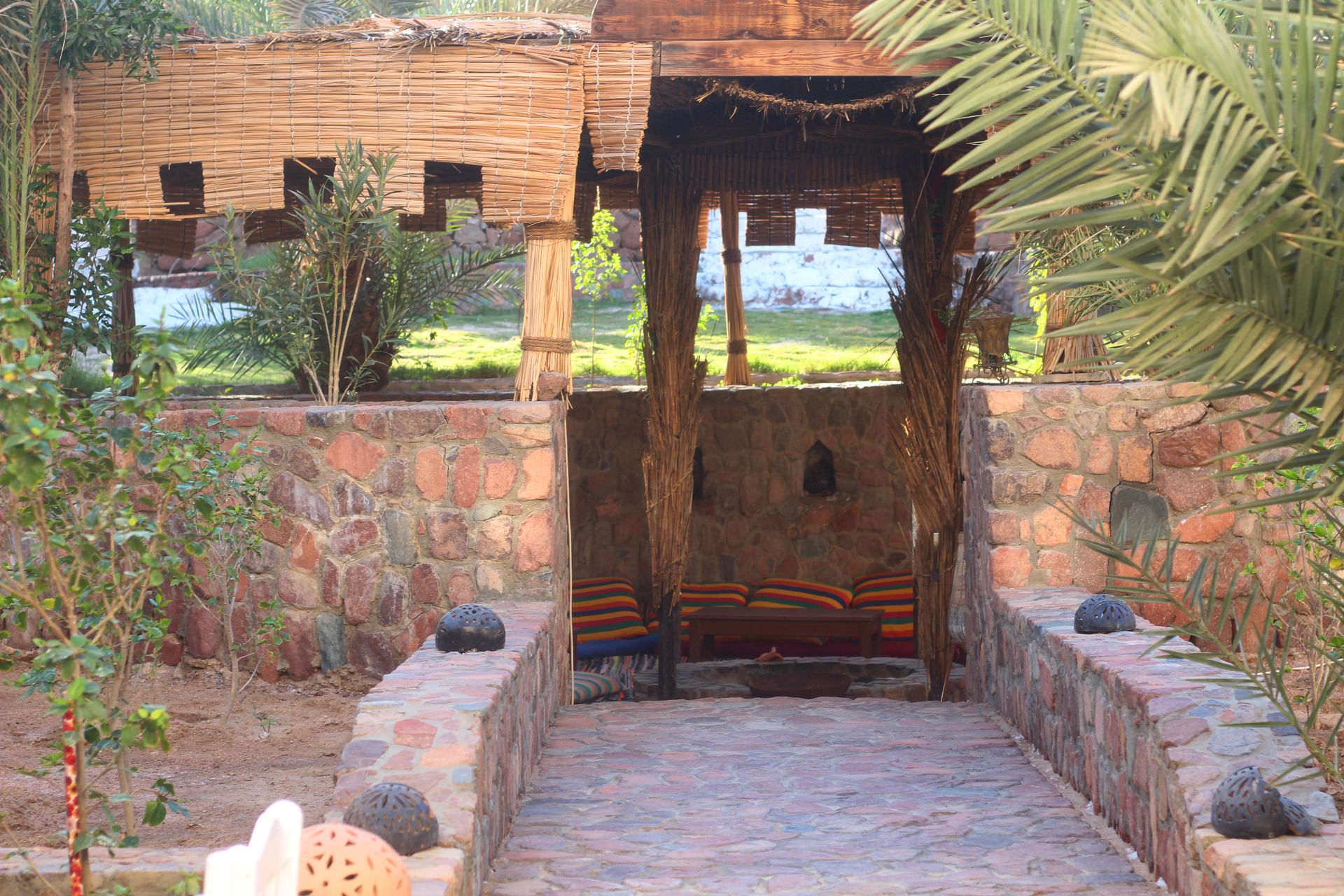 A stone pathway leading to a wooden structure with a thatched roof, surrounded by greenery and a stone wall.