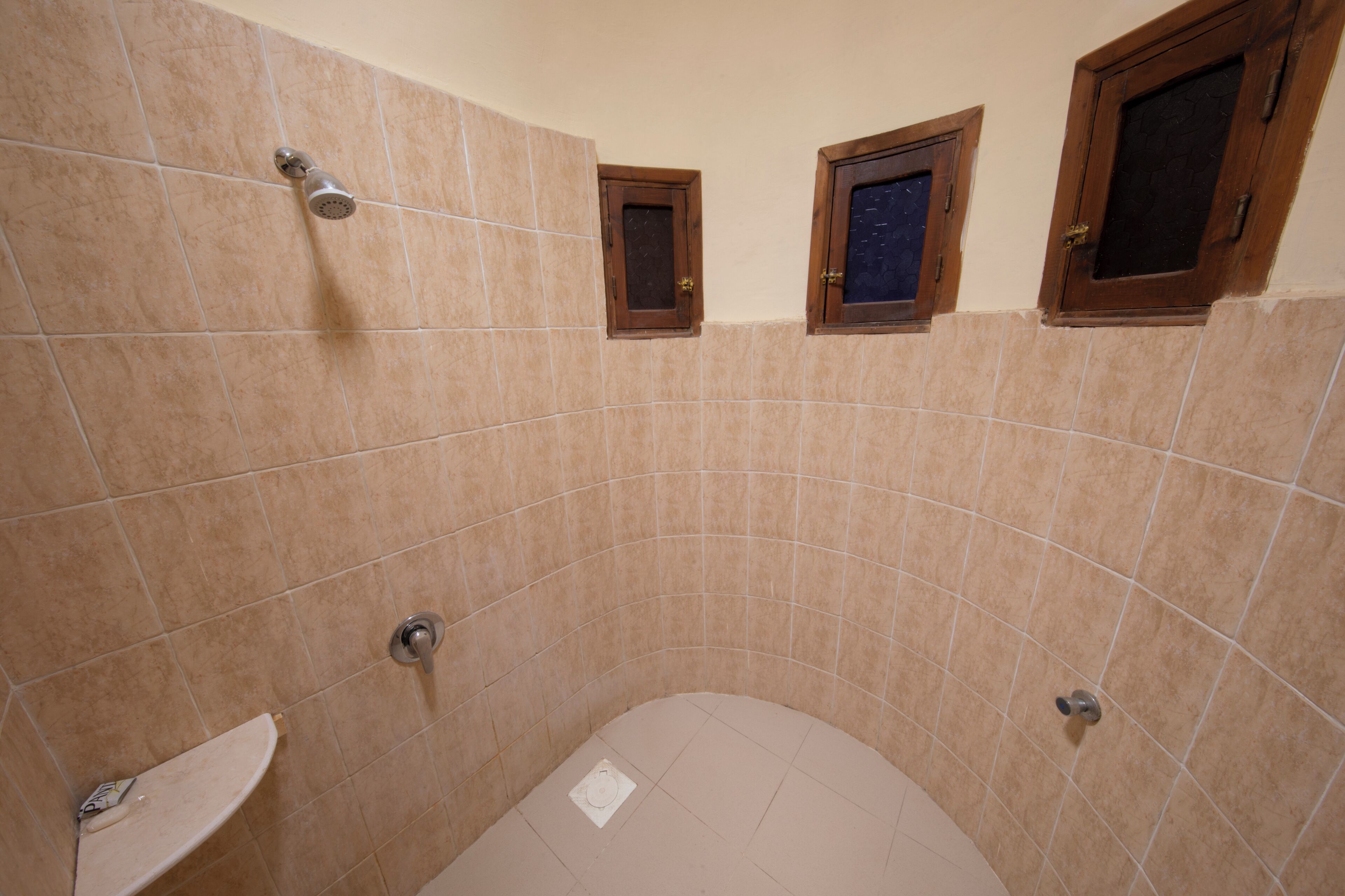 A bathroom with beige tiles, a showerhead, a toilet, and three small windows with wooden frames.