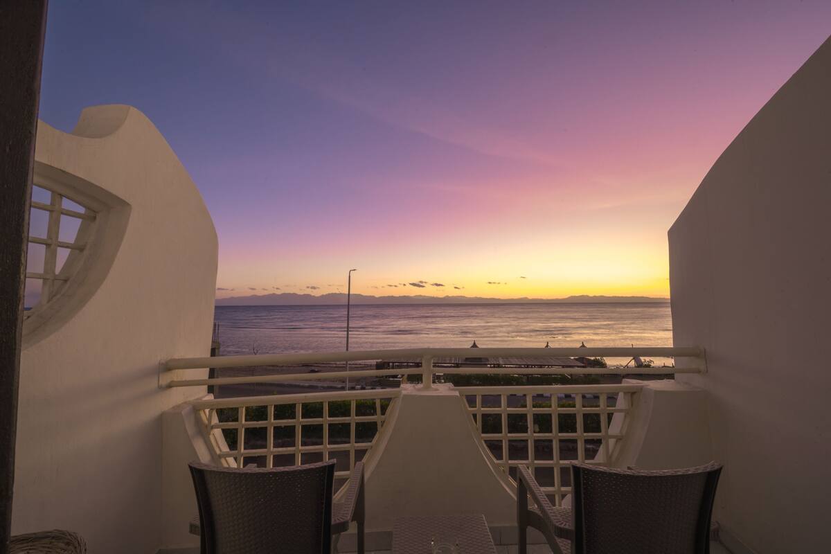 Un balcon avec une table et des chaises, d’où l’on peut admirer le coucher de soleil sur l’océan.